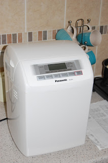 A white Panasonic SD-254 Bread Maker sits on a kitchen worktop near blue mugs hanging on a rack and a sheet of paper. The bread maker features a digital display panel and buttons on the top front.