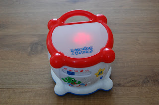 A small, white and red plastic Leapfrog Learning Drum with a handle, a glowing light on top, and colourful designs including a frog and stars, sits on a wooden floor—an engaging educational toy for children.
