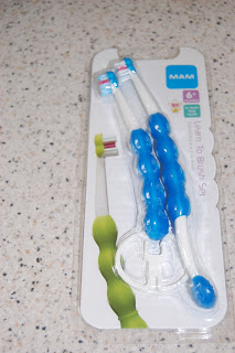A packaged MAM Learn to Brush Set with two blue and white toothbrushes for children aged 6 months and up, displayed on a speckled worktop. This toothbrush set is perfect for early oral care routines.