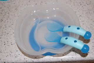 A translucent baby bowl with a blue base sits on a speckled worktop, holding a matching blue baby spoon and fork featuring chunky, dotted handles.