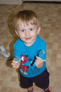 A young child with fair hair, wearing a blue cartoon t-shirt, smiles up at the camera while holding two small baby spoons. The child stands on a tiled floor, and part of an adult's shoe is visible in the background.
