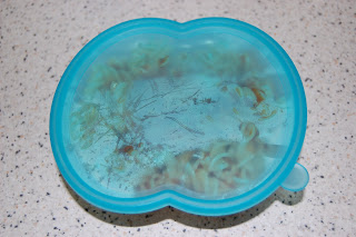 A blue-tinted plastic food container with a foggy lid, showing faint outlines of spiral pasta inside, sits on a speckled worktop next to a baby spoon.