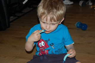 A young boy with blond hair, wearing a blue T-shirt, sits on a wooden floor eating a blue sweet stick, with a blue beaker, toys, and a baby bowl scattered in the background.