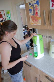 A woman in a black vest uses a green and white SodaStream on a kitchen worktop. Children are visible in the background, with colourful children's artwork hanging on the walls and cupboards.