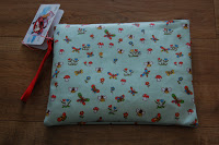 A light blue Pink Lining zippered pouch with a red strap, decorated with small colourful prints of birds, flowers, and mushrooms, lies on a wooden surface. A tag is attached to the zip—perfect as an accessory for any Mum on the Run Bag.