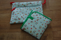 Two fabric pouches with colourful patterns featuring small animals and objects, placed on a wooden surface. Perfect as handy inserts for your Pink Lining nappy bag or Mum on the Run Bag, one is rectangular with green trim, the other has red drawstrings.