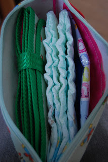 A Pink Lining Mum on the Run Bag holds three folded nappies and a neatly coiled green strap, all arranged side by side inside this stylish changing bag.