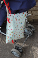 A colourful, patterned Pink Lining Mum on the Run Bag with a red zip hangs on the handle of a pushchair with grey wheels, positioned outdoors on a paved surface near a brick wall.