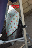 A close-up of a pushchair’s side shows a blue floral-patterned cushion with red and yellow flowers and a red handle, partially placed inside a black mesh pocket, alongside a Pink Lining Mum on the Run Bag. A stone wall and pavement are visible in the background.