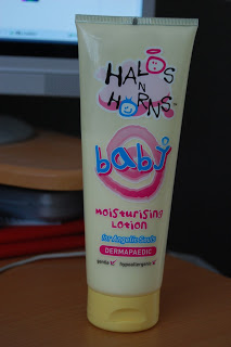 A tube of Halos N Horns Baby Lotion sits on a wooden surface near a computer monitor. The yellow tube with colourful text is labelled as gentle, hypoallergenic, and dermapaedic—perfect for sensitive skin.