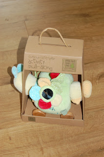 A plush, pastel-coloured Curious Caterpillar pull-along toy packaged in a cardboard box with a handle, sitting on a wooden floor. The box is labelled "activity pull along.