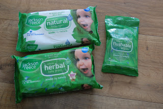 Three packs of Jackson Reece natural wipes in green packaging, labelled "natural" and "herbal," with a smiling baby’s face on two packs and a smaller "flushable wipes" pack, all placed on a wooden surface.