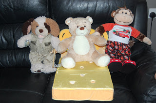 Three stuffed animals—a dog, a bear from Build-A-Bear Workshop, and a monkey—are sitting side by side on a black leather sofa. The bear is in the centre, perched on a yellow butterfly-shaped cushion.
