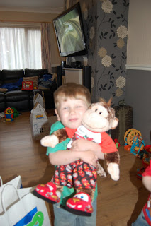 A young boy smiles and hugs a stuffed monkey toy from Build-A-Bear Workshop whilst standing in a living room with toys and shopping bags scattered around. A TV is mounted on the wall in the background.