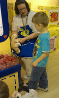 A woman in an apron helps a young boy with a soft toy at Build-A-Bear Workshop. The boy stands near a bin of red items, whilst shelves of boxed toys fill the background.