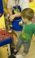 A young child in a green dinosaur shirt watches as an adult in a blue apron stuffs a soft toy at the Build A Bear Workshop station. Red plush hearts are visible in a clear bin nearby, ready for new soft toys.