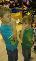 Two young boys stand indoors, each holding a red object to their nose as if pretending to be clowns. Both are wearing blue shirts and jeans, with Build-A-Bear soft toys and toys displayed on shelves in the background.