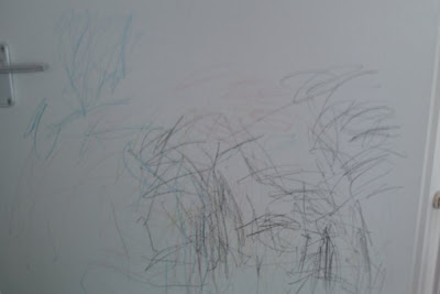 White wall covered with random, colourful scribbles made by crayons or pencils, likely drawn by a child. A silver door handle is visible on the left side—evidence of a little artist’s "My Little Devil" moment.
