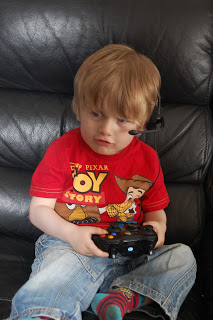 A young boy with light brown hair, wearing a red Toy Story T-shirt, blue jeans, and colourful socks, sits on a black leather sofa holding a video game controller and wearing a gaming headset—perfect for family gaming nights.