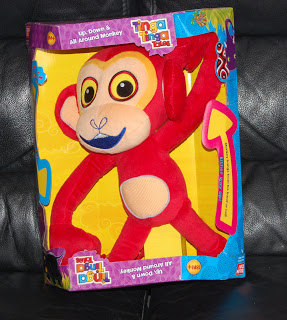 A red plush Monkey with big eyes and blue lips sits in a colourful yellow and purple "Tinga Tinga Tales" box labelled "Up Down & All Around Monkey," displayed on a black leather surface.