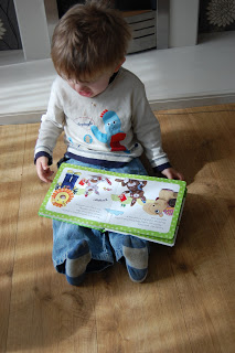 A young child sits on a wooden floor, looking down at an open picture book filled with colourful animal illustrations, wearing a white shirt with a blue dinosaur—ready for their own treasure adventure like Driver Dan's Story Train.
