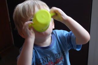 A young child with blond hair is holding and drinking from a bright green, spill-proof beaker, tilting their head back. The child is wearing a blue T-shirt, and their face is mostly hidden by the beaker.