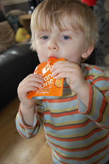 A young child with blond hair wearing a striped shirt drinks from an Orange One food pouch indoors.