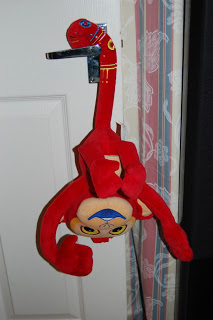A red plush Tinga Tinga Tales monkey toy with long arms is hanging upside down from a door handle, its head and arms dangling below. The white door and nearby patterned curtains complete this playful Up Down All Around scene.