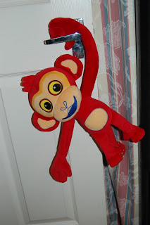 A red plush Monkey toy, inspired by Tinga Tinga Tales, with big yellow eyes hangs by one arm from a silver door handle, next to a white door and patterned curtains.
