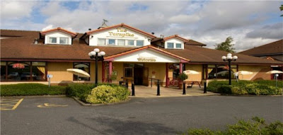 A single-storey building with a sign reading "The Turnpike Brewers Fayre," featuring a brown roof, large windows, outdoor seating, and surrounded by greenery under a partly cloudy sky.