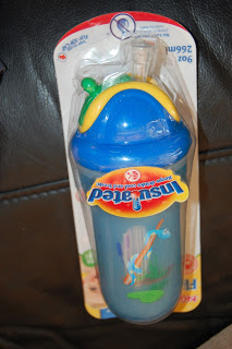 A blue and yellow Nuby Flip-it Cup with a straw, decorated with animal illustrations, is sealed in its original packaging against a dark background.