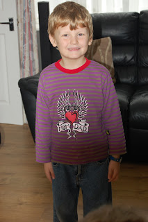 A young boy with light brown hair smiles, standing indoors on a wooden floor. He wears a Rock A Bye Baby Clothing purple and grey striped shirt with a heart and wings graphic that says "I love Dad," and blue jeans. A black sofa is in the background.