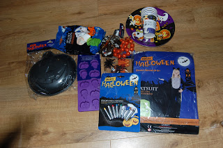 A variety of Halloween-themed products from Aldi are arranged on a wooden floor, including a bat-shaped platter, purple silicone mould, orange beads, a costume, face paint, a plate, and a packet of serviettes.