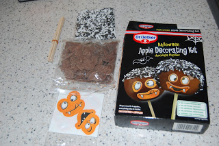 A Halloween Apple Decorating Kit by Dr Oetker featuring chocolate flakes, desiccated coconut, a stick, and two orange monster face stickers—all presented in a box on a speckled worktop.