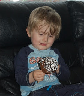 A young child with blonde hair sits on a black leather sofa, smiling and looking at a chocolate cupcake topped with white sprinkles that he decorated using a Halloween decorating kit, holding it proudly in both hands.