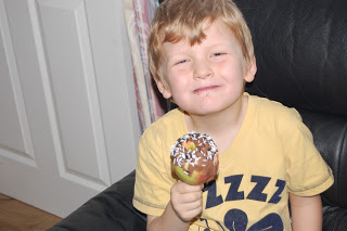 A young boy with fair hair, wearing a yellow T-shirt, smiles whilst holding a chocolate-covered toffee apple made using a Halloween Apple Decorating Kit, sitting on a black chair indoors.