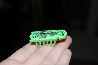 A small green Hexbug Nano toy, resembling an insect with multiple legs, is held in a person's hand against a dark, blurred background.