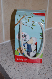 A Wild & Wolf "Charlie and Lola" money box in a colourful gift bag with rope handles, placed on a speckled worktop against a tiled wall.
