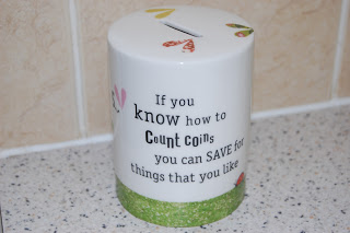 A Charlie and Lola Money Box by Wild & Wolf, featuring colourful leaf designs and the quote: "If you know how to count coins, you can save for things that you like," sits on a speckled worktop against a tiled wall.