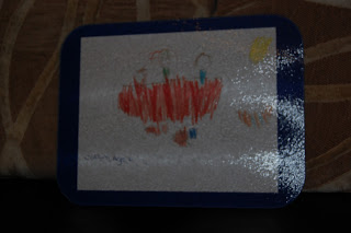 A child’s colourful drawing with red, orange, blue, and yellow shapes on white paper, bordered by a dark blue frame, displayed against a patterned brown background—perfect for art fundraising with Funmats.