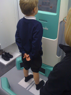 A young boy stands on a platform facing a machine, hands clasped behind his back. Wearing a navy jumper, black shorts, and socks with his Clarks shoes off to the side, he awaits his turn as an adult stands nearby—perhaps it's time for Callum's Clarks review.