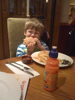 A young boy with blond hair and a striped shirt is sitting at a Brewers Fayre restaurant table, smiling as he holds a large sandwich. There are chips, dip, and a bottle of orange drink on the table in front of him.