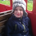 A young child in a blue coat and knitted winter hat with ear flaps sits on a wooden bench inside a red vehicle at Thomas Land, Drayton Manor, with greenery outside—capturing the excitement of a Magical Christmas adventure.