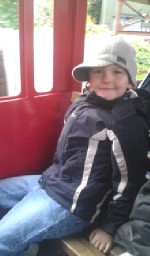 A young child wearing a grey cap, black jacket, and blue jeans sits smiling on a bench inside a red vehicle at Drayton Manor’s Thomas Land, capturing the joy of a magical Christmas adventure.
