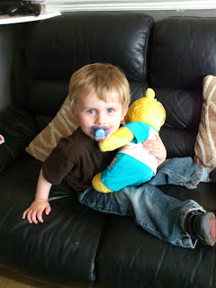 A young child with blond hair and a dummy sits on a black leather sofa, hugging a yellow soft toy from Build A Bear Workshop. Two patterned cushions are behind him, highlighting the joy in supporting children.