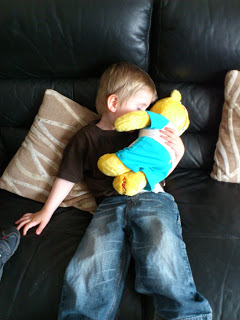 A young child sitting on a black sofa hugs a yellow teddy bear from Build-A-Bear Workshop, wearing a blue shirt. Two beige patterned cushions are behind the child, who is looking away and facing the teddy bear, supporting children with comfort and care.