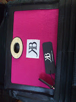 A pink and black bag with a circular gold handle, a white patch featuring the initials "RB," and a matching black tag attached to the zip, also displaying the "RB" logo—perfect for any Kate Benjamin Purse Review.