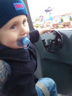 A young child with a blue dummy and blue knitted hat sits on a ride at Thomas Land, holding onto a black steering wheel and looking out with curiosity, ready to enjoy the magic of Drayton Manor's Magical Christmas.