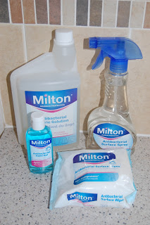 Four Milton Antibacterial products—a liquid bottle, spray bottle, wipes pack, and small bottle—are arranged on a speckled worktop in front of a tiled wall.