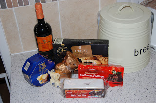 A festive Christmas Hamper of holiday treats sits on the kitchen worktop, featuring Aldi goodies like mince pies, panettone, chocolate rabbit, stollen loaf, Christmas pudding, and a bottle of sherry by a large cream-coloured bread bin.